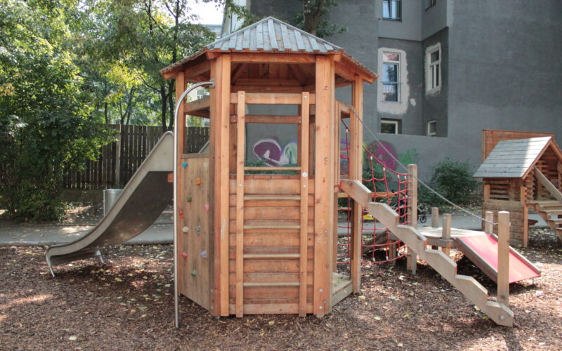 Ein Spielplatz aus Holz mit einer Rutsche, einer Kletterwand, einer Seilbrücke und einer Treppe, der im Freien auf Mulch liegt, mit Bäumen und Gebäuden im Hintergrund.