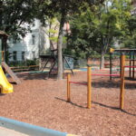 Ein Spielplatz mit einer gelben Rutsche, Klettergerüsten und einem Holzspanbelag, umgeben von Bäumen und nahe gelegenen Gebäuden.