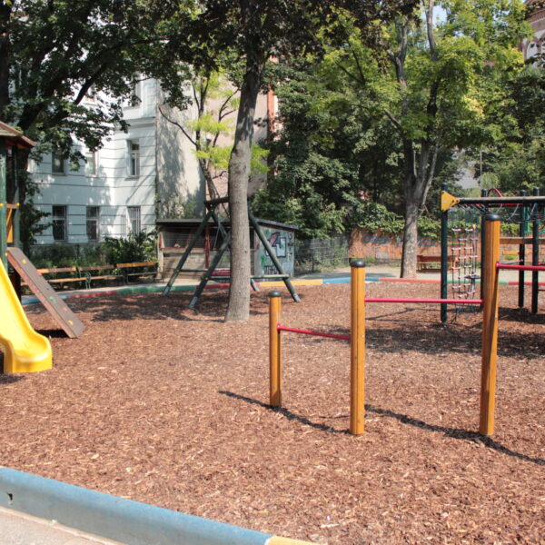 Ein Spielplatz mit einer gelben Rutsche, Klettergerüsten und einem Holzspanbelag, umgeben von Bäumen und nahe gelegenen Gebäuden.
