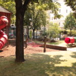 Ein Spielplatz mit einer roten Röhrenrutsche, einem Seilkletternetz und einer roten Tunnelkonstruktion, umgeben von Bäumen und einer Rasenfläche.