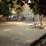 Ein ruhiger Spielplatz im Freien mit Bänken, Picknicktischen, einem Holzspielhaus und einem eingezäunten Sportplatz im Hintergrund.