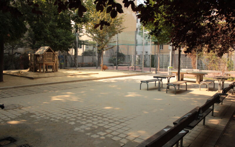 Ein ruhiger Spielplatz im Freien mit Bänken, Picknicktischen, einem Holzspielhaus und einem eingezäunten Sportplatz im Hintergrund.