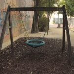 Ein Spielplatz mit einer runden Netzschaukel, die an einem Holzrahmen aufgehängt ist, umgeben von Mulch und mit Graffiti beschmierten Wänden im Hintergrund.