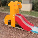 Eine kleine Spielplatzrutsche in Form einer Giraffe, mit orangefarbenen Seiten und einer roten Rutschfläche, die auf einem mit Mulch bedeckten Boden steht.