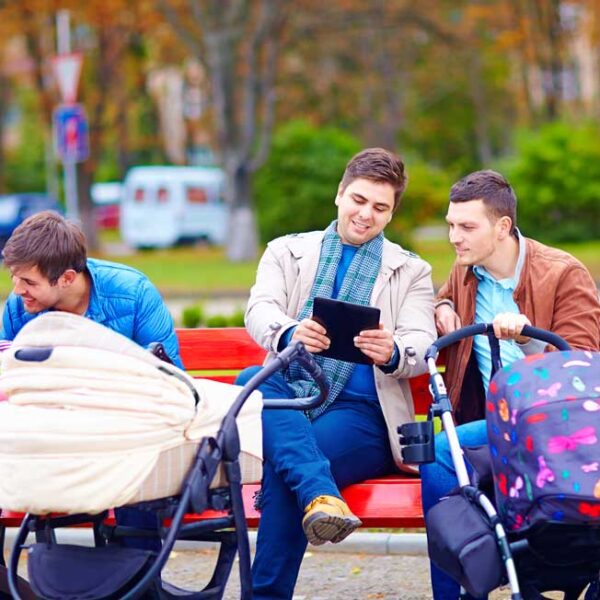 Drei Männer sitzen mit zwei Kinderwagen auf einer roten Parkbank. Einer hält ein Tablet, ein anderer kümmert sich um ein Baby und der dritte hält einen Kinderwagengriff. Im Hintergrund sind Bäume und geparkte Fahrzeuge zu sehen.