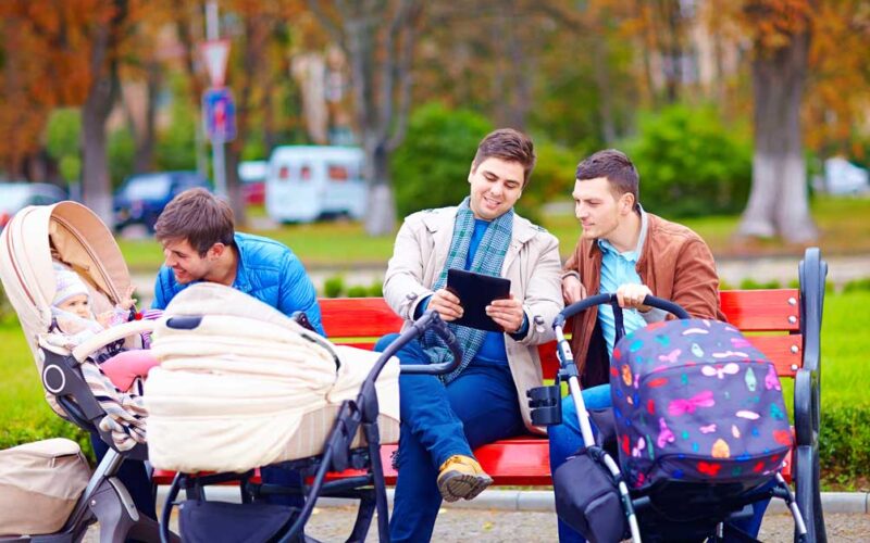 Drei Männer sitzen auf einer Parkbank mit zwei Kinderwagen; ein Mann schaut auf ein Tablet, ein anderer kümmert sich um ein Baby, und der dritte hält einen Kinderwagengriff.