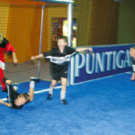 Vier Kinder spielen Hallenfußball; eines in Rot schießt den Ball in die Nähe des Tores, während drei in Schwarz reagieren, wobei eines zu Boden fällt.