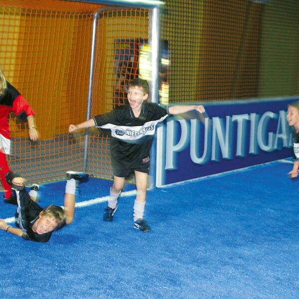 Vier Kinder spielen Hallenfußball; eines in Rot schießt den Ball in die Nähe des Tores, während drei in Schwarz reagieren, wobei eines zu Boden fällt.
