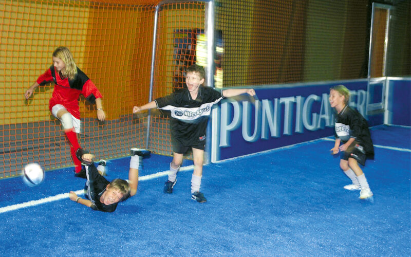 Vier Kinder spielen Hallenfußball; eines in Rot schießt den Ball in die Nähe des Tores, während drei in Schwarz reagieren, wobei eines zu Boden fällt.