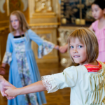 Fünf kleine Kinder, einige davon in historischen Kostümen, bilden in einem reich verzierten Raum mit Golddekor einen Kreis an den Händen.