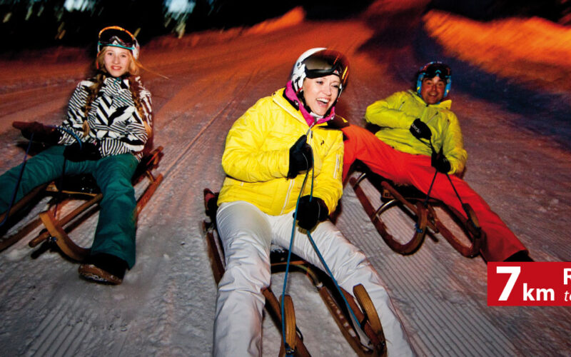 Drei Personen in Winterkleidung fahren nachts auf Holzschlitten einen verschneiten Hang hinunter. In der unteren rechten Ecke steht auf einem Banner „7 km Rodelspaß! Rodelspaß!“.