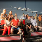 Eine Gruppe von Personen sitzt in einem Fahrsimulator und scheint eine Szene zu erleben, in der ein großes Flugzeug über dem Bildschirm im Hintergrund fliegt.