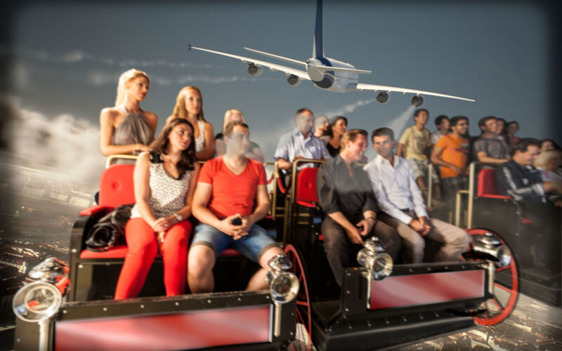 Eine Gruppe von Personen sitzt in einem Fahrsimulator und scheint eine Szene zu erleben, in der ein großes Flugzeug über dem Bildschirm im Hintergrund fliegt.