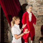 Eine Frau gestikuliert in Richtung einer Wachsfigur, die einen roten Anzug aus dem 18. Jahrhundert trägt, während mehrere Personen in einem schwach beleuchteten Raum mit Steinwänden zusehen.