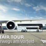 Ein geparktes Flugzeug auf der Rollbahn mit einem VISITAIR TOUR Bus daneben am Flughafen Wien, mit Terminalgebäuden im Hintergrund.