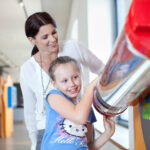 Eine Frau steht hinter einem lächelnden Mädchen, das in einem hell erleuchteten Wissenschaftsmuseum mit einem Exponat interagiert, das eine große rot-silberne Röhre zeigt.