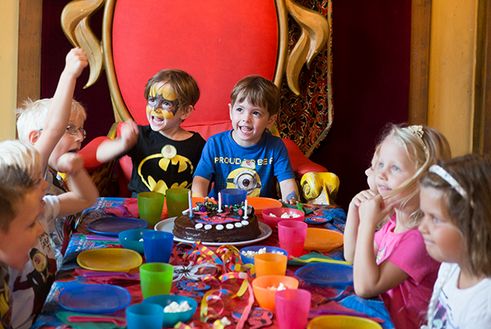 Eine Gruppe kleiner Kinder sitzt um einen geschmückten Tisch mit einem Schokoladengeburtstagskuchen, bunten Tassen und Partydekorationen. Zwei Jungen in der Mitte wirken aufgeregt und lächeln.