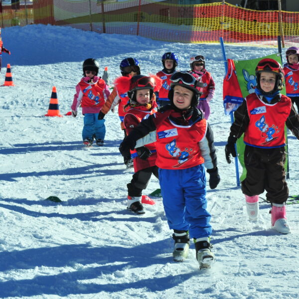 Eine Gruppe von Kindern in Skikleidung und roten Westen läuft in einer Skischule auf dem Schnee, während Lehrer und Sicherheitskegel in der Nähe sind.