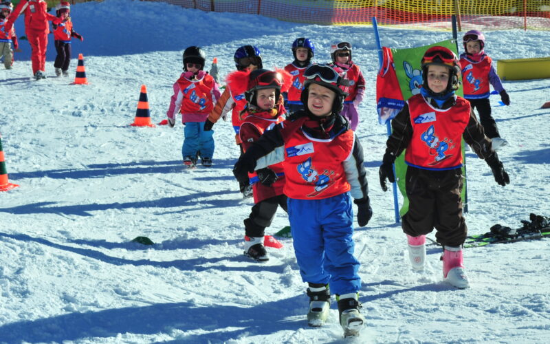 Eine Gruppe von Kindern in Skikleidung und roten Westen läuft in einer Skischule auf dem Schnee, während Lehrer und Sicherheitskegel in der Nähe sind.