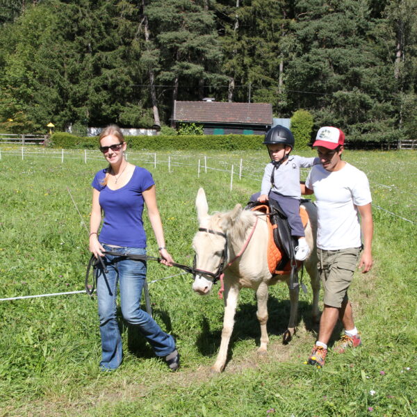 Ein Kind, das einen Helm trägt, reitet auf einem kleinen weißen Pferd, das von zwei Erwachsenen geführt wird, über einen grasbewachsenen Weg in einer ländlichen, bewaldeten Gegend.