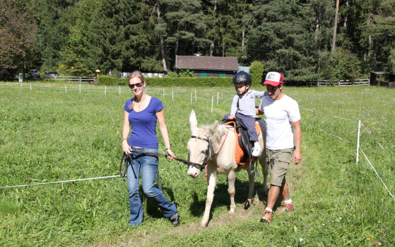 Ein Kind, das einen Helm trägt, reitet auf einem kleinen weißen Pferd, das von zwei Erwachsenen geführt wird, über einen grasbewachsenen Weg in einer ländlichen, bewaldeten Gegend.