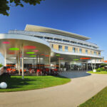 Modernes Gebäude mit geschwungener Architektur, bunten, kreisförmigen Lichtern an der Decke, Rasen und mehreren roten Kinderwagen, die vor dem Eingang geparkt sind, unter einem sonnigen Himmel.