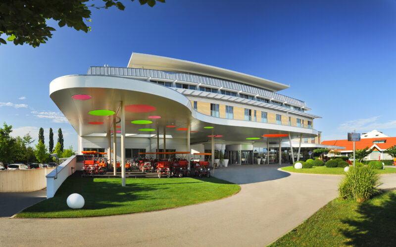 Modernes Gebäude mit geschwungener Architektur, bunten, kreisförmigen Lichtern an der Decke, Rasen und mehreren roten Kinderwagen, die vor dem Eingang geparkt sind, unter einem sonnigen Himmel.