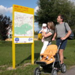 Eine Familie mit Kinderwagen und Baby steht an einem sonnigen Tag in einer Parkanlage neben einem großen gelben Schild mit der Aufschrift „Baby on Tour“.