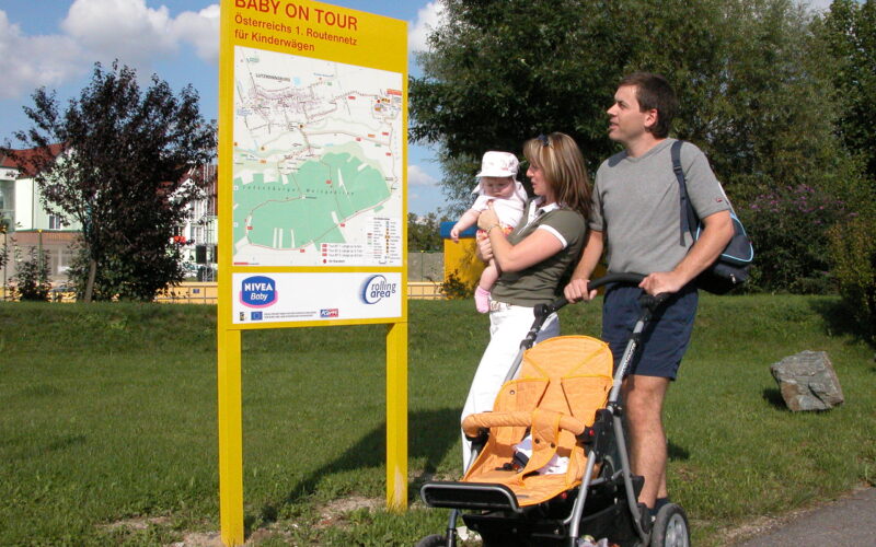 Eine Familie mit Kinderwagen und Baby steht an einem sonnigen Tag in einer Parkanlage neben einem großen gelben Schild mit der Aufschrift „Baby on Tour“.