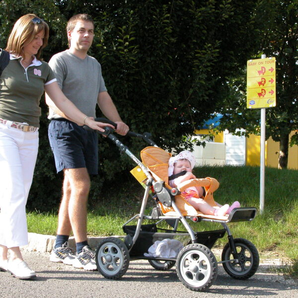 Eine Frau, ein Mann und ein Baby im Kinderwagen gehen an einem sonnigen Tag auf einem gepflasterten Weg in der Nähe eines gelben Schildes und Bäumen spazieren.