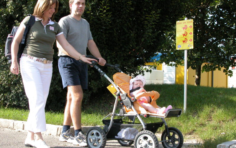 Eine Frau, ein Mann und ein Baby im Kinderwagen gehen an einem sonnigen Tag auf einem gepflasterten Weg in der Nähe eines gelben Schildes und Bäumen spazieren.
