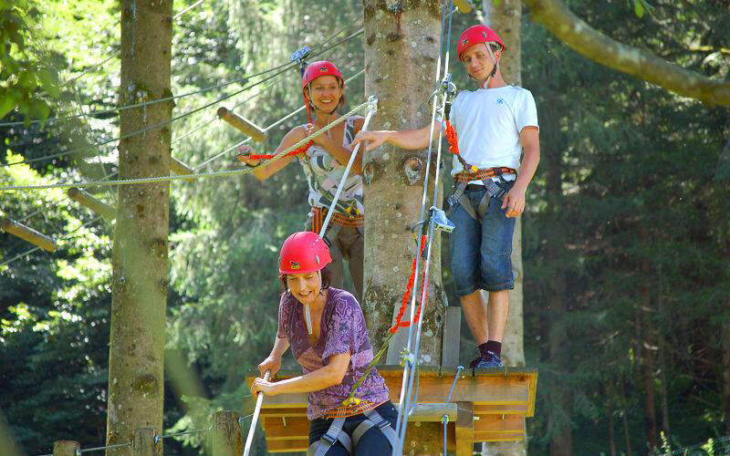 Drei Personen mit Helmen und Gurten stehen auf einer Holzplattform in einem Wald und nehmen an einem Hochseilgarten-Abenteuer teil.