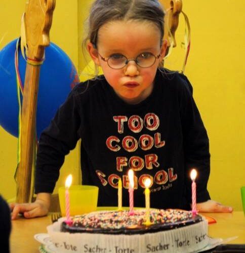 Junges Mädchen mit Brille bläst Kerzen auf einer Geburtstagstorte aus, sie trägt ein "Too Cool for School"-Shirt, im Hintergrund sind Luftballons und Partydekorationen zu sehen.