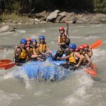 Eine Gruppe von Personen mit Schwimmwesten und Helmen fährt auf einem Fluss Wildwasser-Rafting, hält orangefarbene Paddel und lächelt in die Kamera.