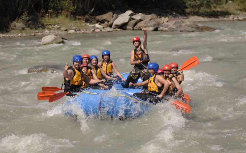 Eine Gruppe von Personen mit Schwimmwesten und Helmen fährt auf einem Fluss Wildwasser-Rafting, hält orangefarbene Paddel und lächelt in die Kamera.