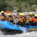 Sechs Personen mit Helmen und Schwimmwesten paddeln auf einem blauen Schlauchboot durch die Stromschnellen eines Flusses.