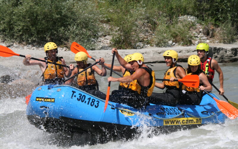 Sechs Personen mit Helmen und Schwimmwesten paddeln auf einem blauen Schlauchboot durch die Stromschnellen eines Flusses.