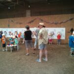 Kinder und Erwachsene nehmen an einer Kunstaktion teil und malen große Dinosaurierzeichnungen an die Wände der Scheune; einige Kinder tragen orangefarbene Hüte, und zwei Erwachsene beobachten die Szene.