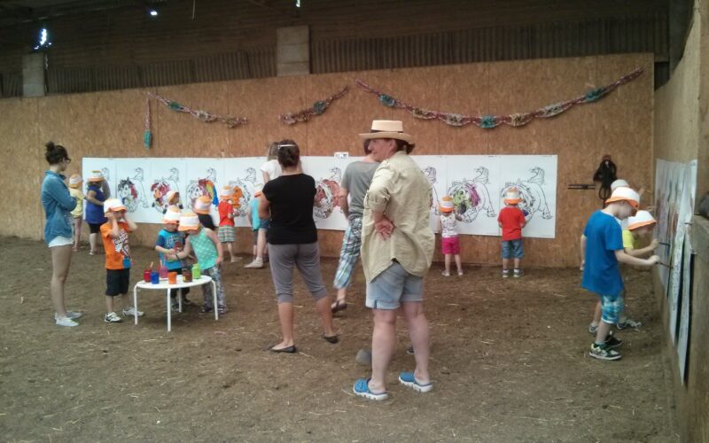 Kinder und Erwachsene nehmen an einer Kunstaktion teil und malen große Dinosaurierzeichnungen an die Wände der Scheune; einige Kinder tragen orangefarbene Hüte, und zwei Erwachsene beobachten die Szene.