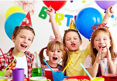 Vier Kinder mit Partyhüten feiern an einem Tisch mit Getränken, Snacks und Luftballons im Hintergrund.