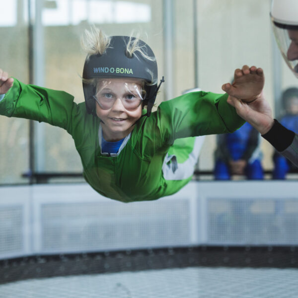 Ein Kind in einem grünen Anzug und Helm schwebt in einem vertikalen Windkanal, während ein Lehrer seinen Arm zur Anleitung hält.