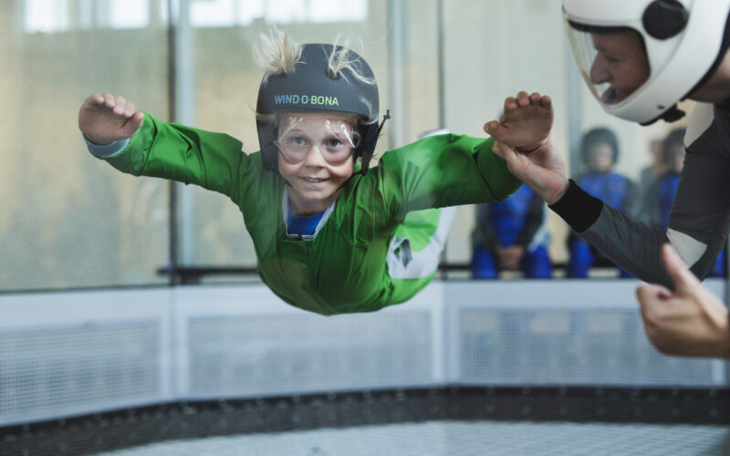 Ein Kind in einem grünen Anzug und Helm schwebt in einem vertikalen Windkanal, während ein Lehrer seinen Arm zur Anleitung hält.
