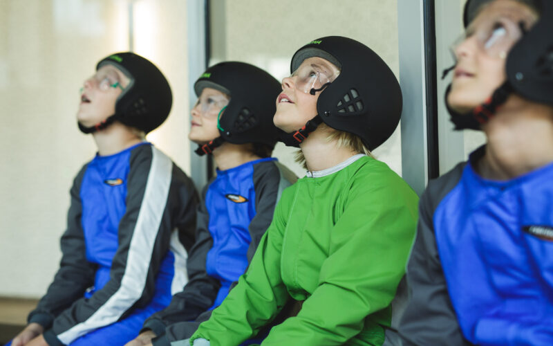 Vier Kinder mit Helmen, Overalls und Schutzbrillen sitzen drinnen und schauen mit aufmerksamen Blicken nach oben.