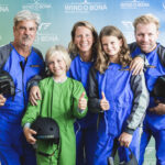 Fünf Personen in Fluganzügen posieren lächelnd und mit erhobenem Daumen für ein Gruppenfoto vor einer Indoor-Skydiving-Kulisse von Windobona.