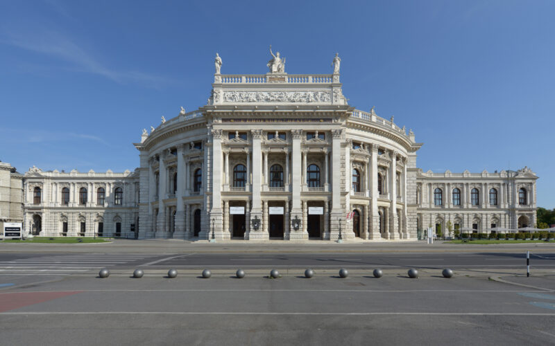 Frontansicht eines großen neoklassizistischen Gebäudes mit Säulen, Statuen auf dem Dach und kunstvollen architektonischen Details vor einem klaren blauen Himmel.