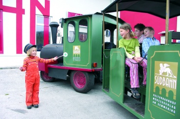 Ein Kind in orangefarbener Latzhose und mit Hut pustet Seifenblasen in der Nähe von drei Kindern, die in einem grünen SüdBahn-Minizug sitzen.