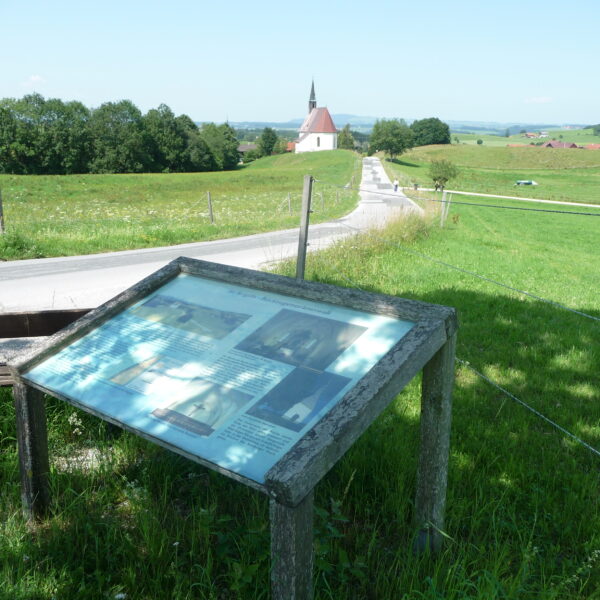 Informationsschild an einer Landstraße mit einer Kirche und Feldern im Hintergrund an einem sonnigen Tag.