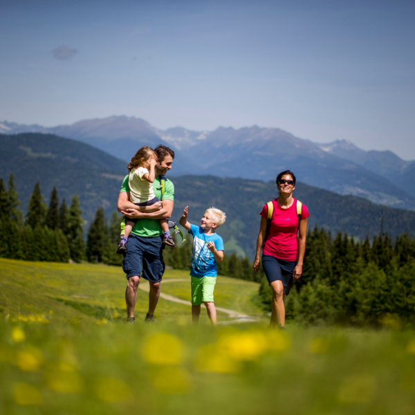 Eine vierköpfige Familie wandert unter klarem Himmel durch eine Graswiese mit Bergen und Bäumen im Hintergrund.