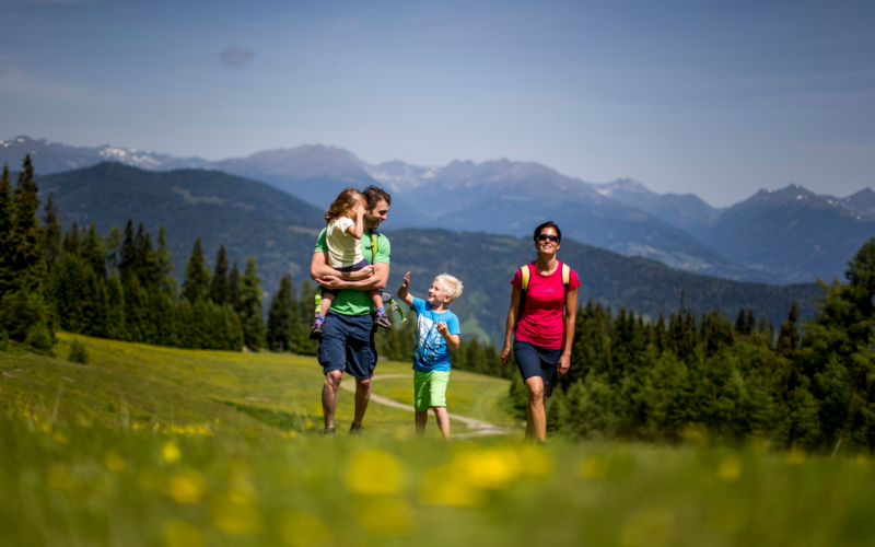 Eine vierköpfige Familie wandert unter klarem Himmel durch eine Graswiese mit Bergen und Bäumen im Hintergrund.