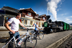 Zwei Personen mit Fahrrädern stehen auf einem Bahnsteig neben einem alten Dampfzug, mit Holzhäusern und blauem Himmel im Hintergrund.